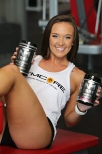 A fit woman in a gym smiling and holding fitness supplements, showcasing active lifestyle.