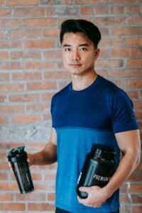 Asian man in a blue t-shirt holding two supplement bottles, standing against a brick wall.