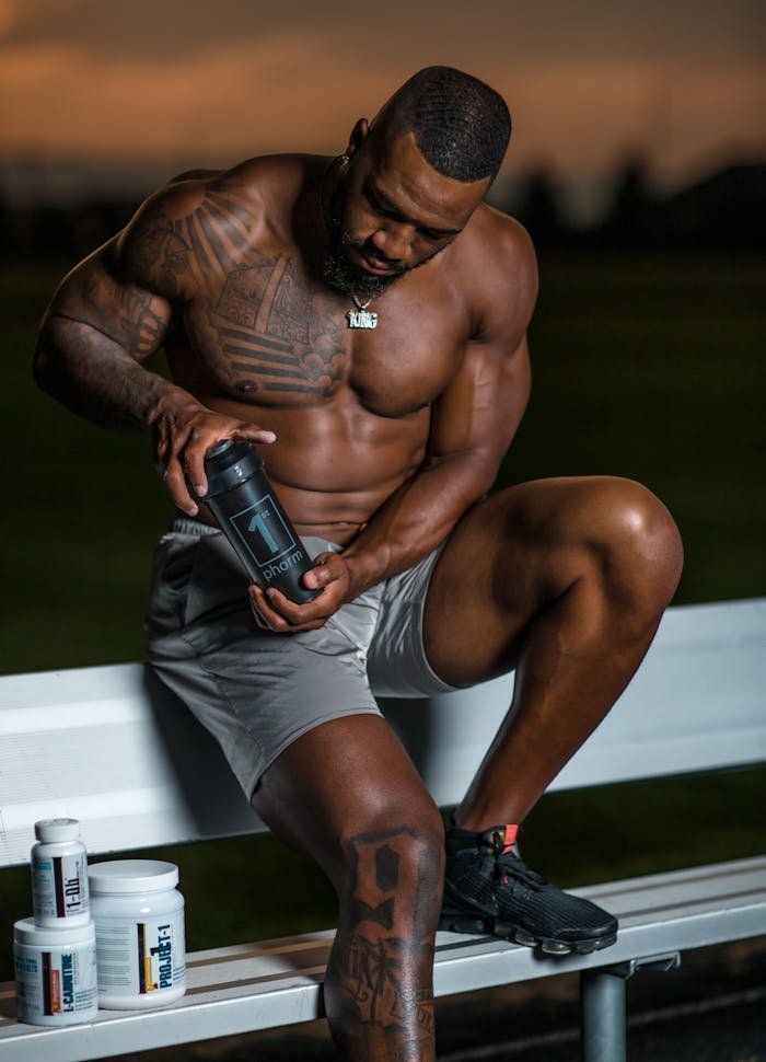 Tattooed bodybuilder seated outdoors with supplements, emphasizing fitness and health.