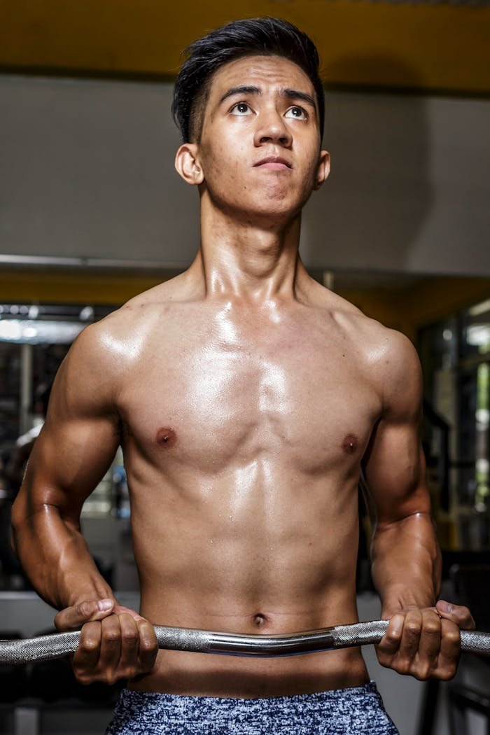 Shirtless young man lifting weights in a gym, showcasing strong biceps and muscular build.